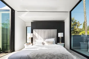 A modern residential bedroom with white walls, a black panel headboard, and white bedding.