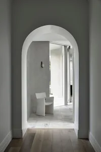 A minimalist lounge area with a white upholstered chair on a marble floor, viewed through a white archway.