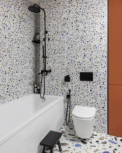 Modern bathroom interior with white terrazzo tiles, white bathtub, white toilet, black fixtures, and wooden storage cabinet.