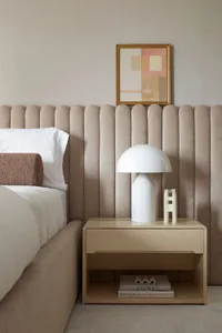 Minimalist bedroom with a beige velvet upholstered bed, white bedding, and a sleek white table lamp on a wooden nightstand.