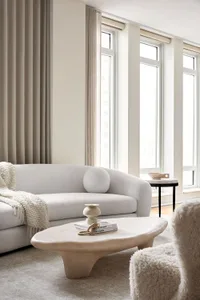 A modern, neutral-toned living room with a white sofa, marble coffee table, and large windows.