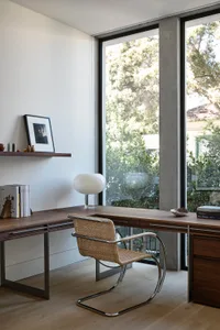 Modern home office featuring a wooden desk, minimalist chair, and large window with views of greenery.