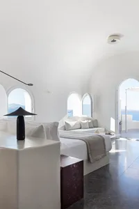 Modern, minimalist bedroom with white walls and concrete floor, featuring a cozy bed and seating area with arched windows.
