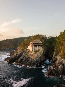 A modern, minimalist cliffside house with a thatched-roof terrace overlooking the ocean.