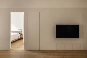 A minimalist apartment with a living room featuring a wooden floor, a television mounted on a wall, and access to a bedroom w