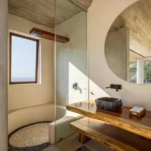 Modern minimalist bathroom featuring sea view through a large window, with a concrete wall, wooden vanity, and stone sink.