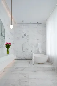 A luxurious modern bathroom featuring white marble walls and floor tiles, a freestanding bathtub, and a minimalist design.