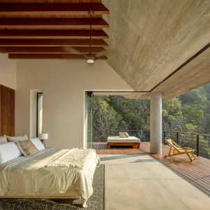 A modern bedroom featuring a concrete ceiling and wooden beams, with a view of lush greenery outside through large windows.