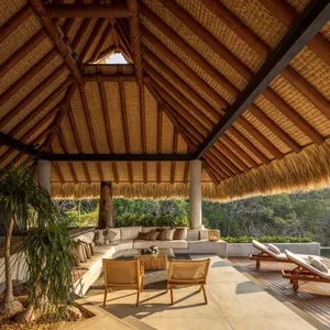Modern outdoor lounge featuring curved sofas, wicker chairs, and a thatched roof, surrounded by lush greenery.