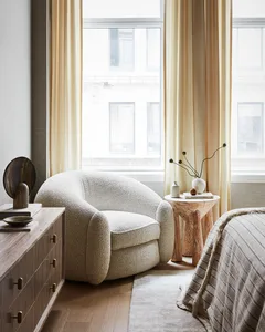 A cozy, modern bedroom with a textured white chair, wooden dresser, and beige curtains by a window.