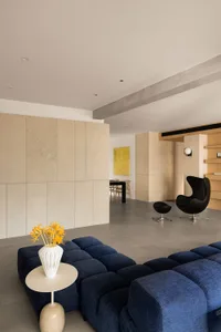 A modern living room with a concrete floor, featuring a blue sofa, a white side table, and a black chair with a footrest.