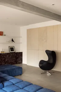 Modern living room with blue sectional sofa, black armchair, marble accent wall, concrete ceiling beam, and tiled floor.