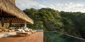A luxurious outdoor living area with a thatched roof, wooden deck, pool, and lush green surroundings.