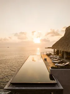 A luxurious outdoor pool area overlooking the ocean at sunset, featuring wooden decks, thatched roofs, and concrete accents.