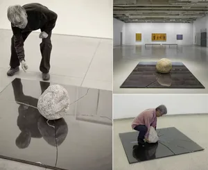 A reflective metal floor displays a large spherical stone, with a person interacting with it in a museum.