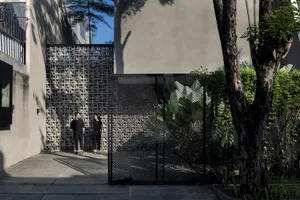 A modern courtyard with concrete walls, a black mesh fence, and lush greenery, featuring two people standing.