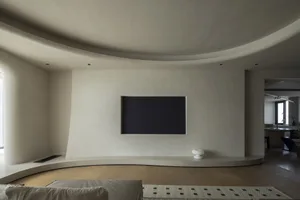 A minimalist living room with a concrete wall and a black flat-screen TV on a curved concrete platform. The floor is made of 