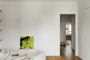 An image of an empty minimalist bedroom with white walls, wood flooring, and a green and yellow framed artwork.