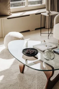 A minimalist living room featuring a glass coffee table with cherries, books, and a vase with dried grasses.