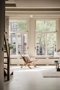 A minimalist living room featuring a wooden chair with a cushion, large windows, and a concrete floor.
