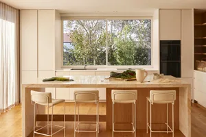 A modern kitchen with a marble countertop, stools, a kitchen island, and a large window overlooking greenery.