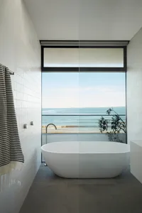 Modern minimalist bathroom featuring a freestanding bathtub with a view of the ocean, tiled walls, and a large window.