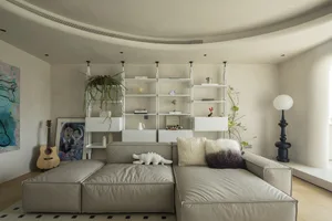 A minimalist living room featuring neutral-colored furnishings, a white bookshelf, and green plants.