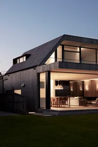A modern house with black siding and large glass windows illuminated against a twilight sky.