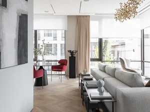 A modern, neutral-toned living room with a large window, white walls, light-colored wooden floor, and minimalist furniture.