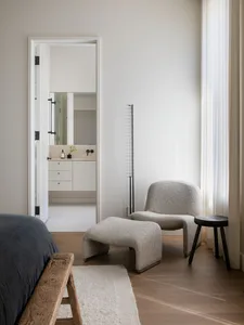 Modern bedroom featuring a neutral color palette with a wooden bench, a plush grey chair, and a minimalist bathroom seen thro