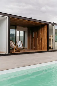 Modern hut with a pool, wooden and glass walls, and two deck chairs.