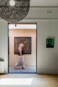 A blurred figure walks through a modern entryway with a wood-paneled wall and abstract painting.