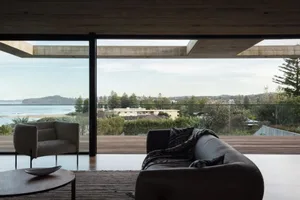 Minimalist living room featuring glass walls and a sea view, with a sofa and armchair, and a wooden floor.