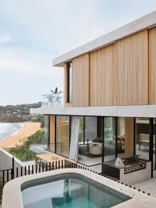 Modern residential exterior with glass walls, wooden slats, and a swimming pool overlooking a beach.