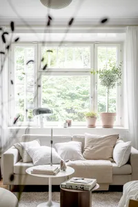 White couch, beige cushions, potted plants, candles, white curtains, and large window