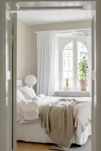 A minimalist bedroom with white textiles and a beige blanket draped over the bed, featuring a large white curtain by the wind
