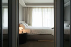 A minimalist bedroom featuring a white fabric bed with soft lighting and light-colored wooden furniture.