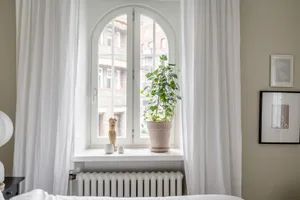 A minimalist bedroom featuring white curtains, a white radiator, and a white window frame with a view of another building thr