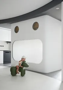A child sits on a green, plastic toy horse in a minimalist play area with white walls and floor.