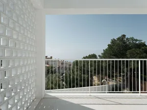 A residential balcony featuring white glass blocks and a metal railing under a clear blue sky and surrounded by greenery.