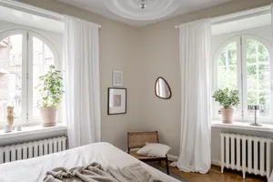 A minimalist bedroom with white curtains and walls, featuring a bed with a soft blanket, a small table with a lamp, and potte