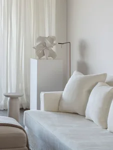 A minimalist living room with a white textured fabric sofa, white curtains, and a modern white sculpture on a pedestal.