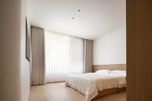Minimalist bedroom with a bed, wooden furniture, light textiles, plastic blinds, white walls, and a wooden floor.