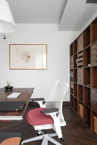 A minimalist office workspace featuring a wooden desk, a red and white ergonomic chair, and a large bookshelf filled with fil