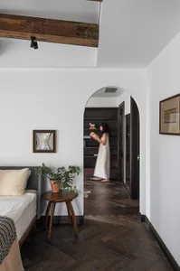 A minimalist bedroom featuring dark wooden flooring, a bed with neutral bedding, a side table with a plant, and a woman stand