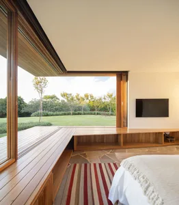 A minimalist bedroom featuring wooden flooring, a large window overlooking a green garden, and a bed with a white blanket.