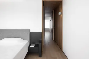 A modern bedroom with a wooden floor, white walls, and a grey upholstered bed. The design features a minimalist aesthetic wit