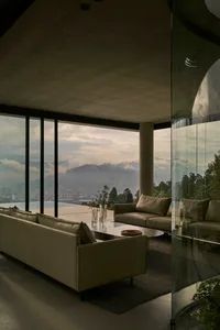 A modern living room with panoramic views of mountains and a cityscape through large glass windows, featuring concrete walls 