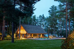 A modern country house with a thatched roof and wooden exterior, surrounded by tall trees and featuring a pool.