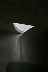 Circular concrete structure with light filtering through, casting vertical shadows on the wall.